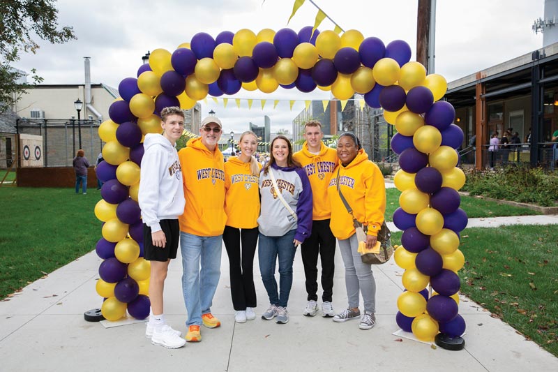 a group of peopole in WCU gear smile for a picture under a purple and yellow balloon arch