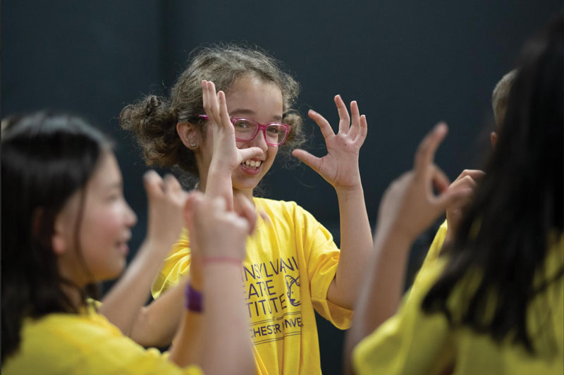 A young girl wearing glasses and a yellow tshirt is smiling in a circle and making motions with her hands