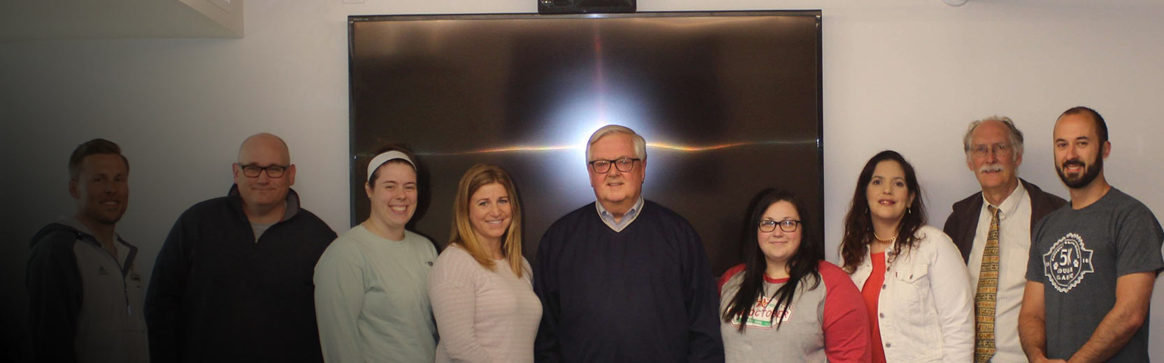 Nine adults stand together indoors in front of a large screen, posing for a group photo.